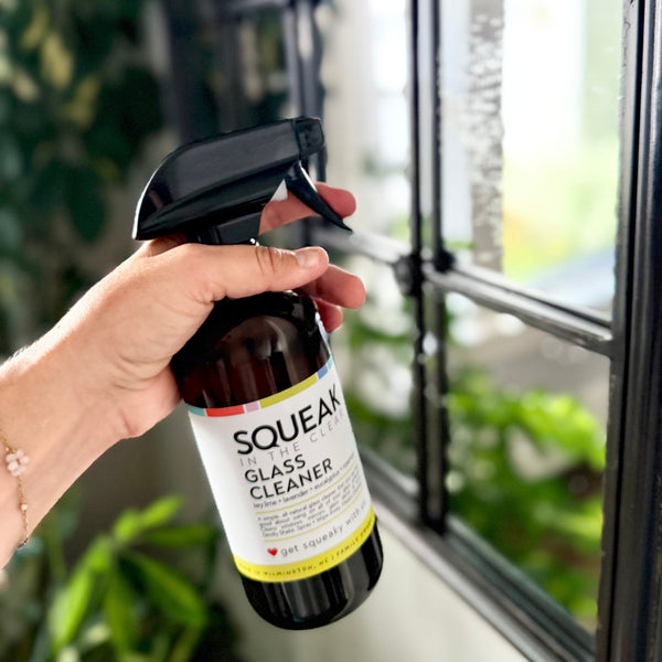 A hand holding a spray bottle of Squeak Glass Cleaner in front of a window with a blurred indoor background.